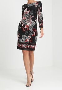 Svart knälång klänning med röda och vita blommiga paisleymönster, trekvartsärmar, bärs med svarta högklackade sandaler.