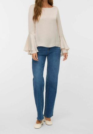 Femme portant un chemisier crème à manches évasées, un jean bleu droit et des chaussures crème à talons bas, debout devant un fond uni.