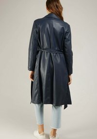 Trench-coat en simili cuir marine avec une taille ceinturée, des manches longues et un col classique. Porté par-dessus un pantalon cropped en denim clair et des baskets blanches.
