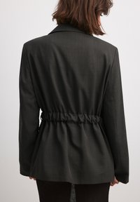 Black, tailored blazer with a structured design, featuring a cinched waist. Made of textured fabric with subtle patterning. Long sleeves.