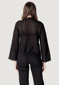 Blouse en crochet noir avec des manches évasées, un ourlet festonné et un design en maille ouverte. Portée avec un jean noir, mettant en valeur une texture superposée.