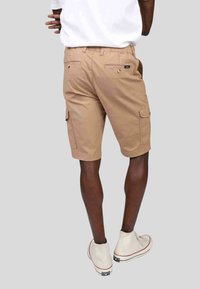 Shorts cargo beiges avec poches latérales, fabriqués en coton durable. Présentent une coupe décontractée et des détails cousus. Portés avec des baskets blanches.