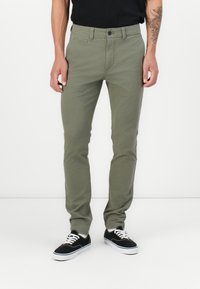 Pantalons en coton verts avec une coupe slim et fermeture à bouton, dotés de poches avant et d'une texture lisse. Associés à des baskets noires.