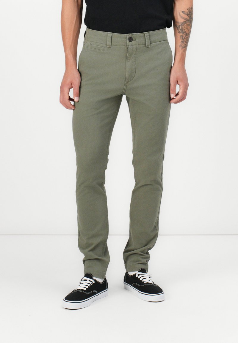 Pantalons en coton verts avec une coupe slim et fermeture à bouton, dotés de poches avant et d'une texture lisse. Associés à des baskets noires.
