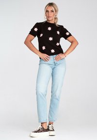 Kobieta w czarnej bluzce z krótkim rękawem ozdobionej różowymi kwiatami, w jasnoniebieskich dżinsach i trampkach na platformie w panterkę, stoi z rękami na biodrach.