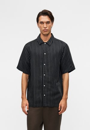 Mladý muž v čiernej krátkym rukávom košeli na gombíky s bielymi vertikálnymi prúžkami, stojaci pred jednoduchým svetlým pozadím.