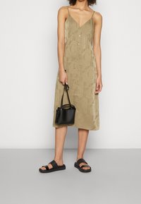 Khaki midi šaty s výstřihem do V a knoflíkovým zapínáním vpředu, s jemným vzorem loga. V kombinaci s černými plochými sandály a malou černou kabelkou.