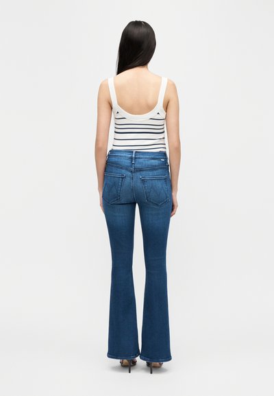 Γυναίκα όρθια με την πλάτη, φορώντας ένα άσπρο τοπ με μαύρες ρίγες και μπλε παντελόνι καμπάνα με ψηλά τακούνια.