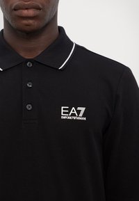 Čierna polokošeľa vyrobená z bavlny, s bielym vyšitým logom na ľavej hrudi a bielym lemovaním na golieri. Troj-buttonové zapínanie.