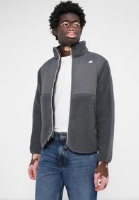 Veste en polaire grise avec un col haut, tissu gris contrasté et un logo blanc sur la poitrine, associée à un jean bleu et une chemise blanche.