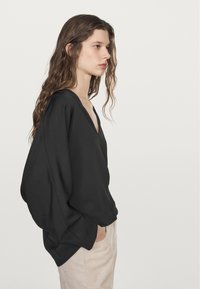 Maglione leggero nero con scollo a V profondo, maniche oversize, texture liscia e vestibilità rilassata, abbinato a pantaloni beige chiaro.