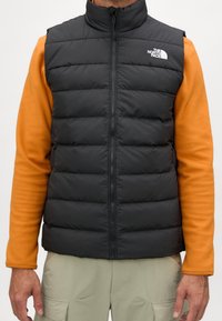 Czarna kamizelka puchowa z wysokim kołnierzem, pikowanym wzorem i zamkiem z przodu. Nosi się ją na pomarańczowym swetrze z logo po lewej stronie.