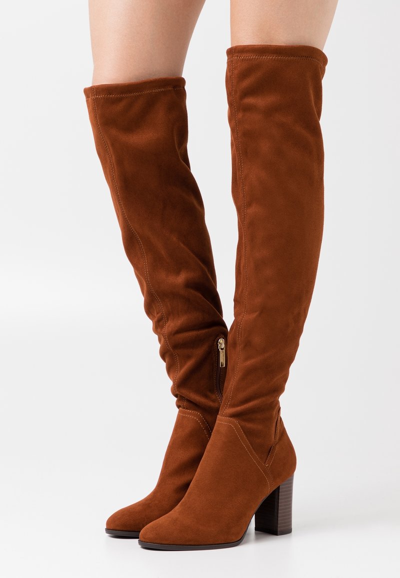 Bottes marron en daim au-dessus du genou avec fermeture éclair latérale et talons blocs portées sur jambes nues sur un fond uni.