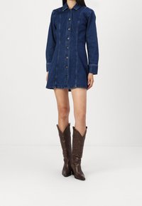 Vestido de camisa de mezclilla, azul oscuro, de manga larga, con cierre de botones en la parte delantera, costuras en contraste. Combinado con botas de vaquero marrones con patrones intrincados.