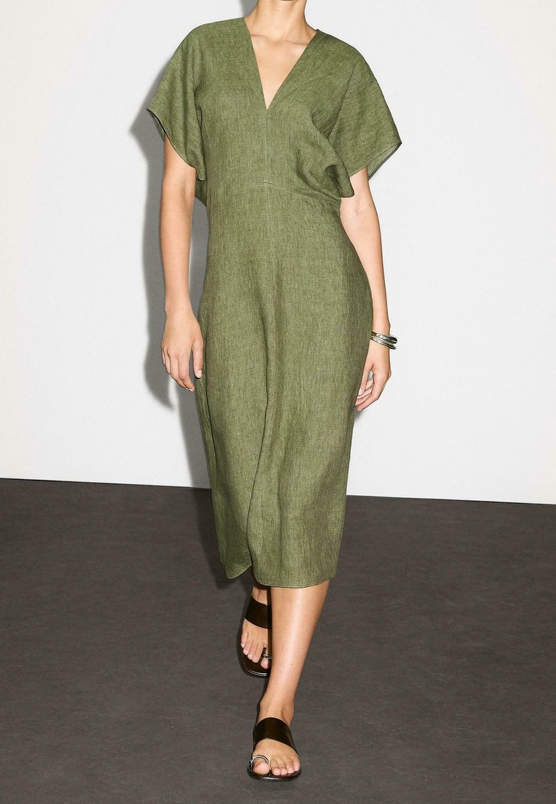 Femme portant une robe vert olive mi-longue à manches courtes et des sandales plates noires, marchant sur un sol sombre contre un mur blanc.