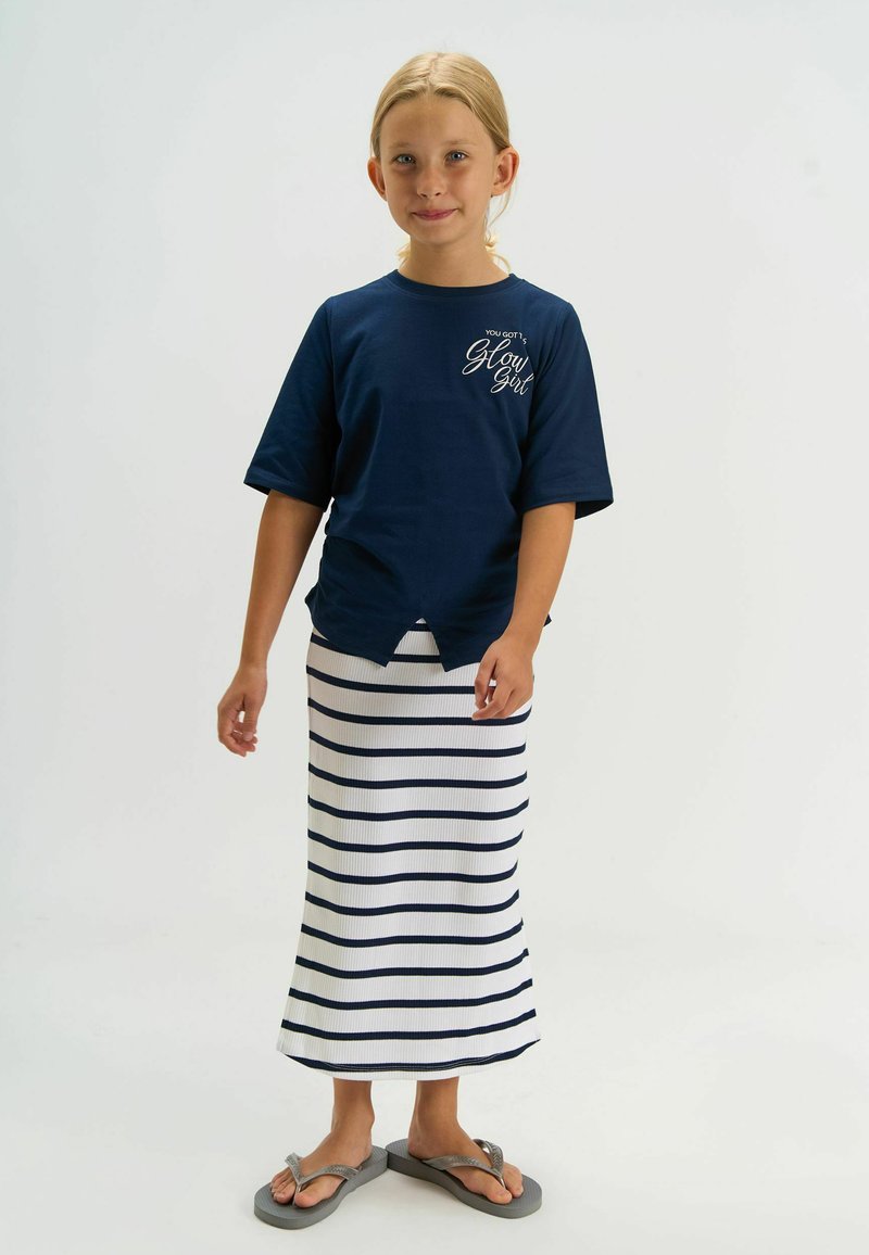 T-shirt bleu marine à manches courtes avec le texte "Glow Girl", associée à une jupe maxi rayée blanche. La fille porte des tongs grises. Design minimaliste, tenue décontractée.