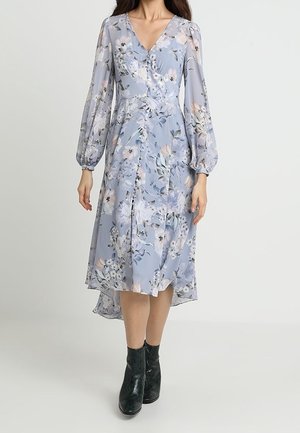 Frau trägt ein hellblaues, geblümtes Midikleid mit langen Puffärmeln und schwarzen Stiefeletten und steht vor einem einfarbigen Hintergrund.