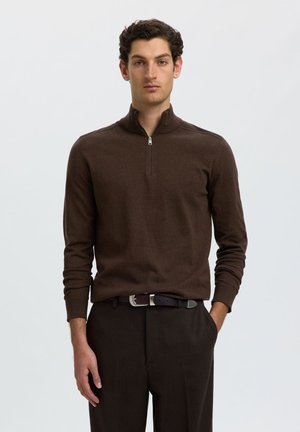Homme aux cheveux bouclés et foncés portant un pull brun à manches longues avec une fermeture éclair au quart et un pantalon brun assorti avec une ceinture noire.