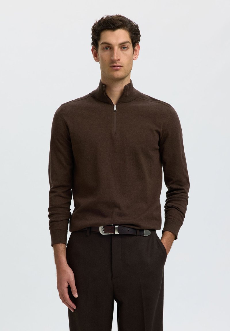 Homme aux cheveux bouclés et foncés portant un pull brun à manches longues avec une fermeture éclair au quart et un pantalon brun assorti avec une ceinture noire.
