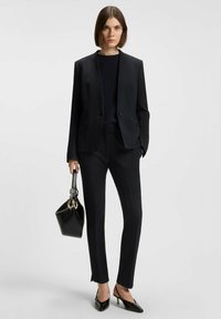 Mørk, struktureret dragt bestående af en tætsiddende blazer og slanke bukser, parret med en sort håndtaske og spidse hæle, alt sammen i en monochrom palet.