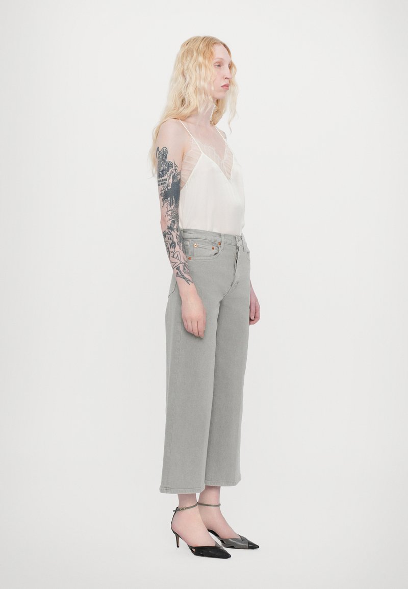 Light gray high-waisted jeans with wide legs, paired with a sheer white top featuring lace accents. Black pointed-toe heels with ankle straps.