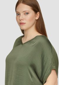 Olive zielona bluzka z dekoltem w serek, wykonana z lekkiego, gładkiego materiału; ma podwinięte krótkie rękawy i luźny krój, eksponując subtelną strukturę.