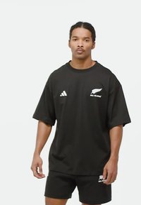 Černé tričko s krátkým rukávem, kulatým výstřihem a volným střihem, se bílými logy Adidas a grafikou "All Blacks" na hrudi.