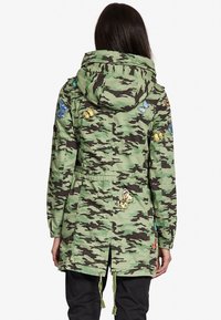Kamouflaged jacka i grönt och svart, med fjärilsbroderier i olika färger. Inkluderar huva och justerbar dragsko i midjan.