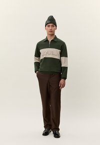 Sötétzöld és bézs színű, "Les Deux" feliratos pulóvert viselő férfi, barna nadrágban, fekete cipőben és sötétzöld sapkában, egyszínű háttér előtt áll.