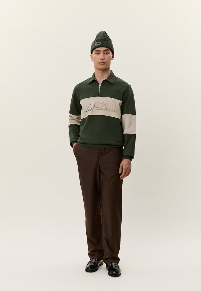 Sötétzöld és bézs színű, "Les Deux" feliratos pulóvert viselő férfi, barna nadrágban, fekete cipőben és sötétzöld sapkában, egyszínű háttér előtt áll.