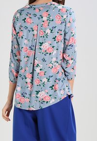 Blusa floreale in azzurro chiaro, con rose rosa e accenti viola. Maniche a tre quarti con dettagli plissettati nella parte posteriore. Tessuto morbido.