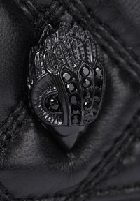 Szczegół czarnej skórzanej torebki na ramię z teksturowanym czarnym metalowym zapięciem w kształcie ptasiej głowy, zdobionym czarnymi kamieniami szlachetnymi i misternymi wzorami.
