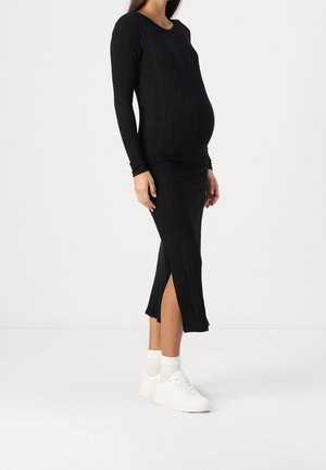 Gravid kvinde iført en lang sort ribstrikket kjole med sidelomme, hvide sokker og hvide sneakers stående på en ensfarvet hvid baggrund.