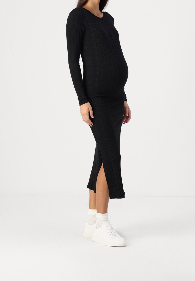 Femme enceinte portant une longue robe noire côtelée avec une fente latérale, des chaussettes blanches et des baskets blanches, debout sur un fond blanc uni.