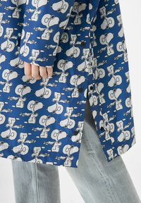 Blauwe waterdichte jas met een herhalend wit cartoonpatroon. Hij heeft een rechte snit, zwarte knopen en lange mouwen. Spijkerbroek zichtbaar.