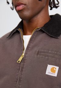 Brun zip-jakke med en kordfløjskrave. Har en sidelomme og et hvidt patch-logo med et orange design. Gyldent lynlåsdetalje.