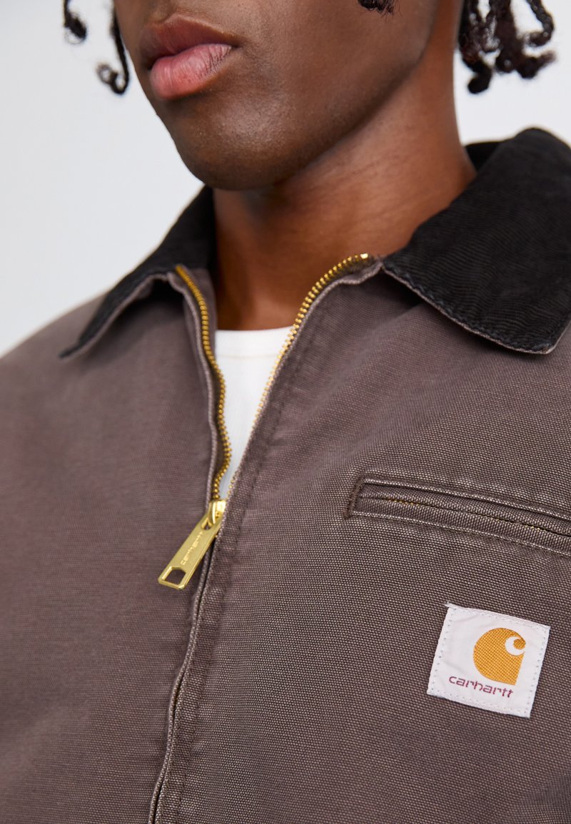 Bruine zip-up jack met een corduroy kraag. Heeft een zijzak en een witte patch logo met een oranje ontwerp. Gouden ritsaccent.