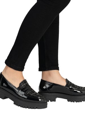 Sorte loafers i laklæder med tekstureret stofdetalje, båret med sorte tætsiddende bukser, vist fra siden i midt-lægniveau.