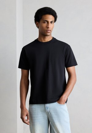 Bărbat tânăr cu păr scurt și creț stă sprijinit de un perete simplu, purtând un tricou negru cu mâneci scurte și blugi bleu deschis, cu mâna în buzunar.