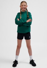 Grøn langærmet sportstop med høj krave, kombineret med sorte shorts. Har hvide detaljer og logo på shortsene. Båret med hvide sokker og sorte sko.