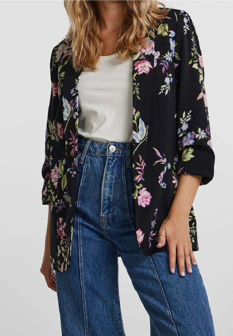 Mujer con blazer floral negro sobre una camiseta blanca y jeans azules de tiro alto, de pie con una mano apoyada en el muslo.