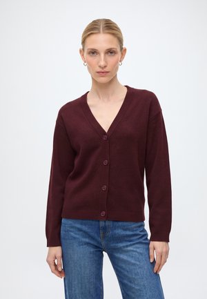 Vrouw met blond haar die gouden oorringen draagt, een donkerrode, knoopjesvest en een blauwe spijkerbroek, staand tegen een effen witte achtergrond.