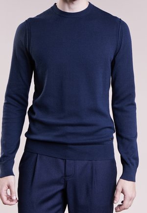 Mann trägt einen enganliegenden marineblauen Pullover mit Rundhalsausschnitt und passende Hose vor schlichtem Hintergrund.