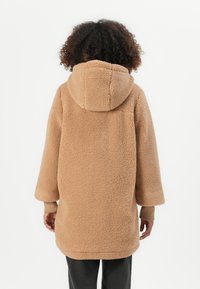 Beige Sherpa-Mantel mit Kapuze, der mit überschnittenen Schultern und weiten Ärmeln gestaltet ist. Weicher Stoff, knielang, mit gerippten Bündchen und Seitentaschen.