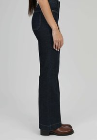 Vue de côté d'une personne portant un jean taille haute bleu foncé et des bottines en cuir marron, sur fond uni.