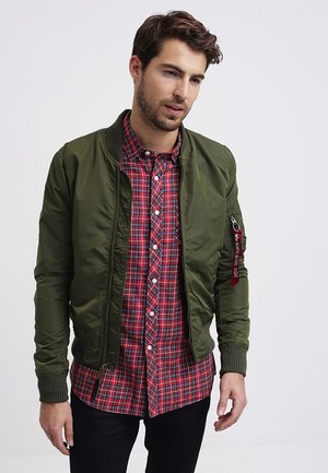 Olive green bomber jacket with a ribbed collar and cuffs, worn over a red and navy plaid shirt. Features zippered pocket and tag detail.
