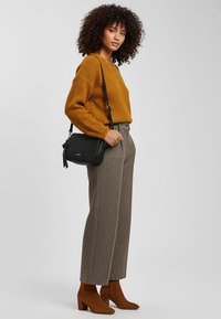 Maglione lavorato a maglia color senape, pantaloni ampi a gamba larga con motivo, stivali marroni alla caviglia e borsa a tracolla nera. Modello posato su uno sfondo neutro.