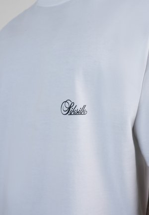 Baltas medvilninis marškinėlis su mažuoju juodu kursyvu parašytu „Siksilk“ logotipu kairėje krūtinės pusėje, parodytas nuo peties iki vidurio liemens.