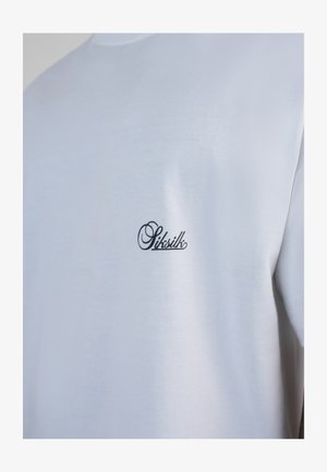 T-shirt en coton blanc avec petit logo "Siksilk" noir en cursive sur la poitrine gauche, porté par une personne visible de l'épaule au milieu du torse.