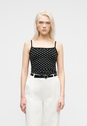 Kvinde med blondt hår iført en sort polkaprikket camisole-top, hvide bukser med høj talje og et sort bælte med sølvspænde, stående oprejst.